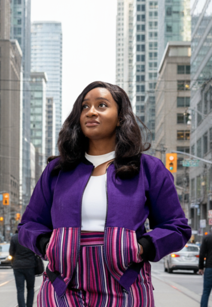 Woman in a purple asomoke bomber jacket and striped pants standing on a city street.